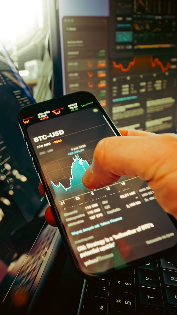A hand holds a smartphone displaying BTC-USD chart amidst digital trading screens, highlighting crypto finance.